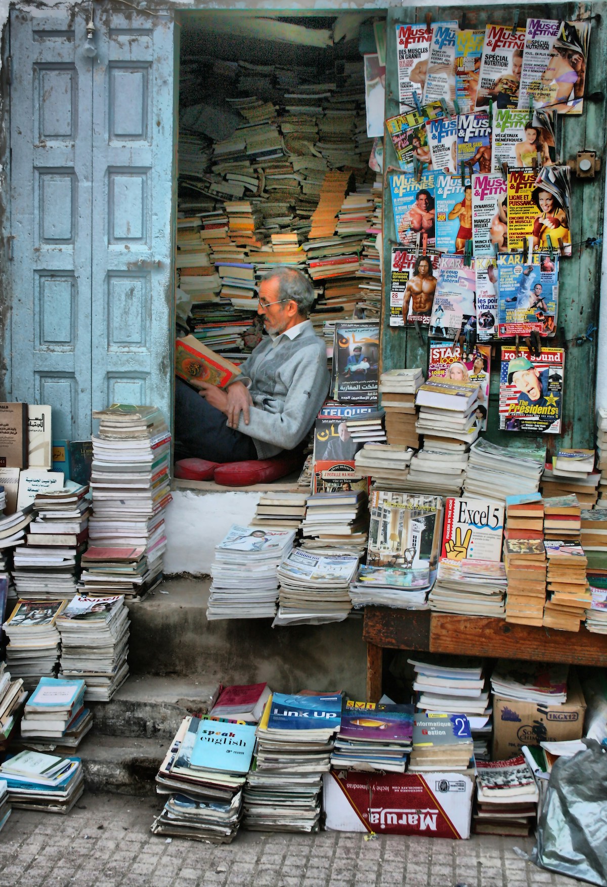 Elderly man reading book surrounded by stacked books and magazines in small shop