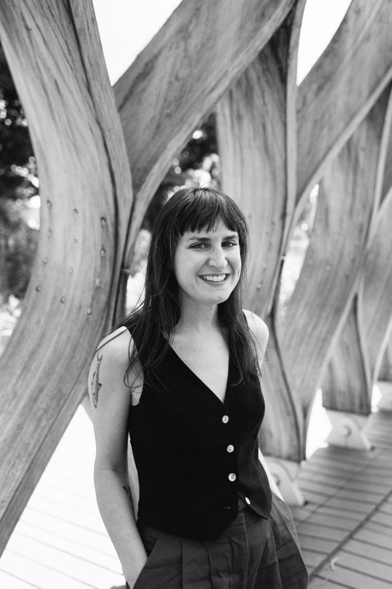 Maggie Summer smiling in black vest top between wooden architectural structures