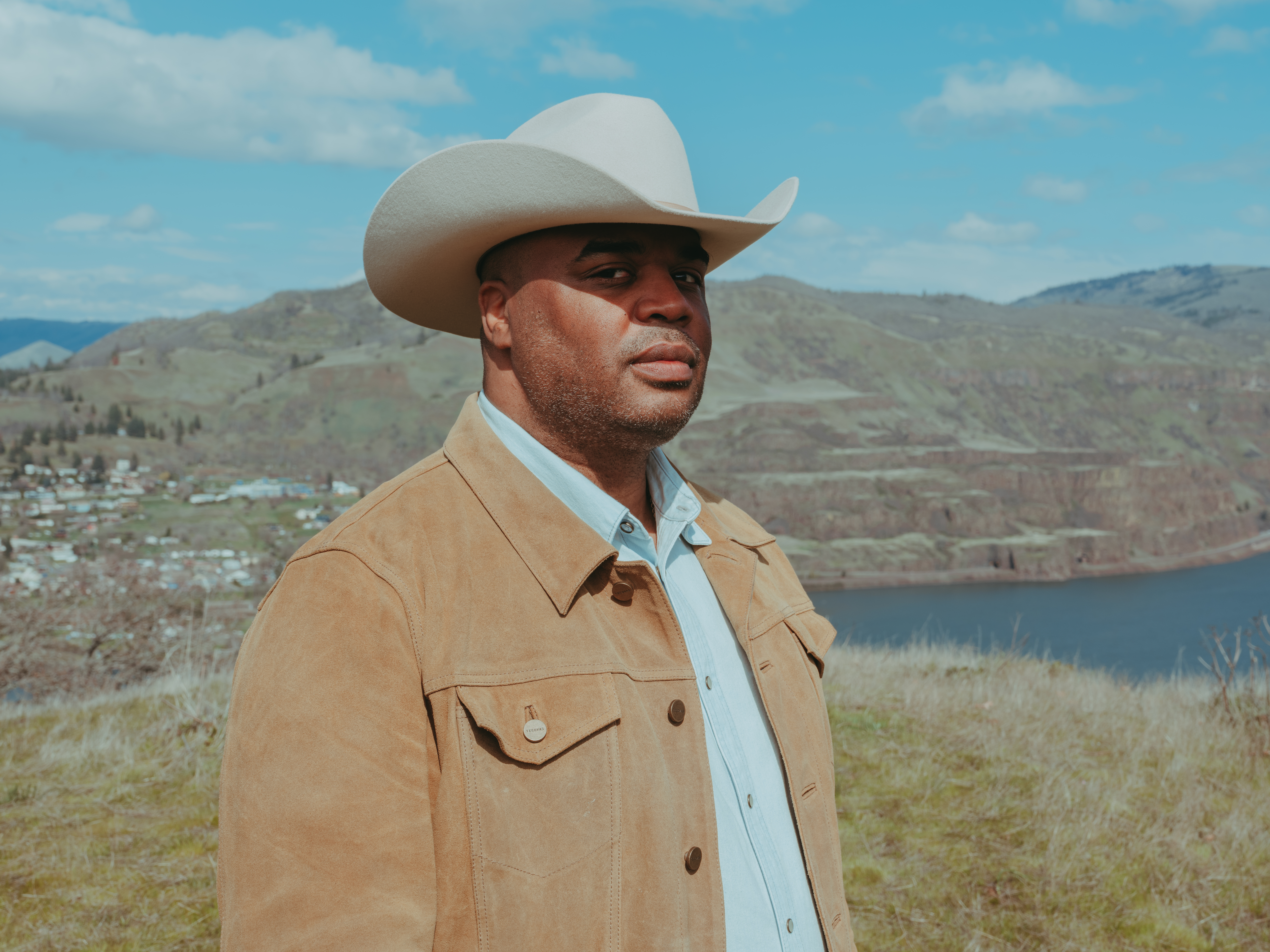 Image of a Black man, wearing a cowboy hat and tan jacket, standing in front of a landscape with a mountain and a lake