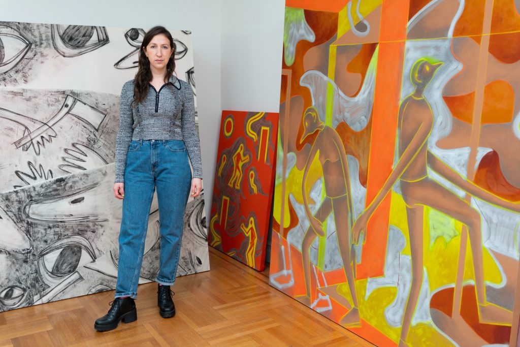 Photo of artist and gallerist Jaqueline Cedar, in front of brightly colored paintings against a wall