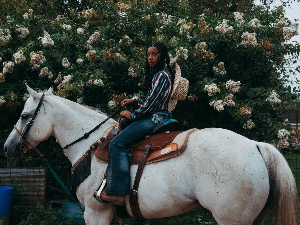 Image of a young Black girl, on a white horse, wearing a floral shirt in front of a floral bush