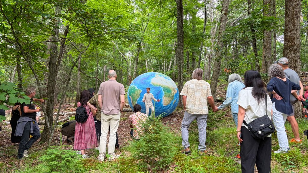 Photo of group of people in a forest watching a performance art piece with a large rendition of a planet Earth