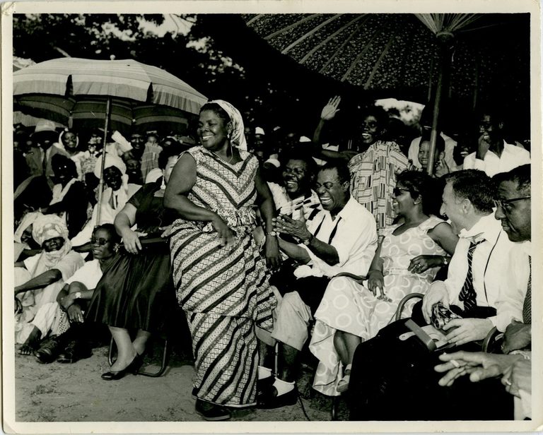 Black and white photo of Louis Armstrong in Ghana, sitting amongst a crowd of people and smiling.